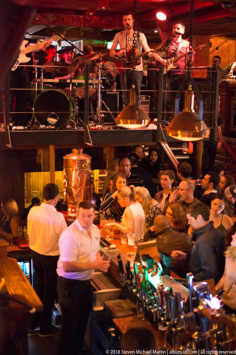 Busy pub bar beneath an elevated live-music stage, with patrons, staff, hanging lamps, and musicians.