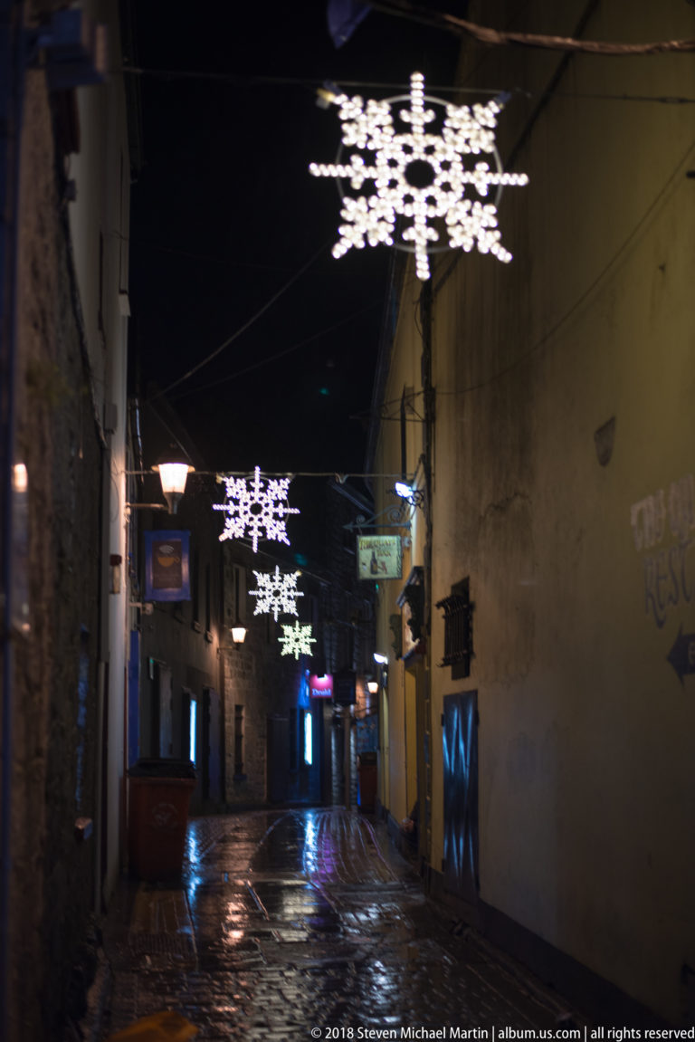 Narrow wet Galway lane at night with large illuminated snowflake decorations suspended overhead.