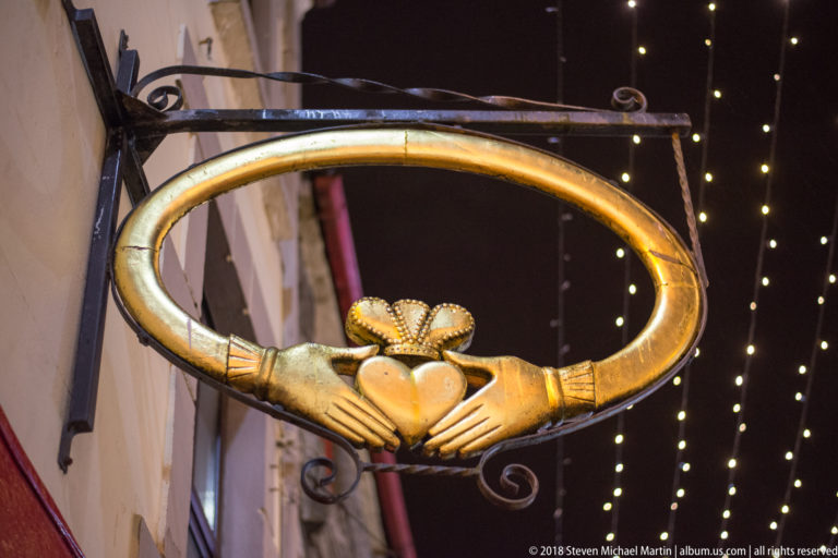 Gold hanging sign shaped as a Claddagh ring, with hands holding a crowned heart against nighttime lights.