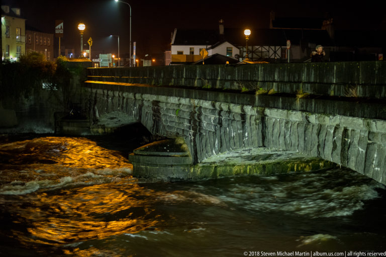Stone bridge with central pier over fast-moving amber-lit river water at night, with buildings beyond.