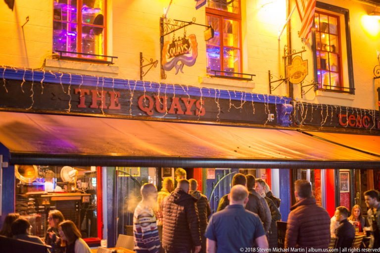 The Quays pub in Galway at night, with its name across the façade and patrons gathered beneath the awning.