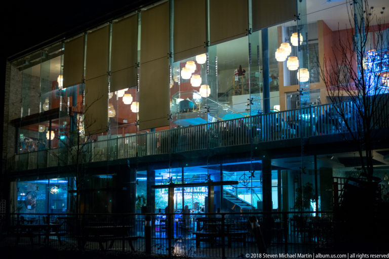 Modern glass building at night with visible stairs, balconies, pendant lights, and blue-lit ground floor.
