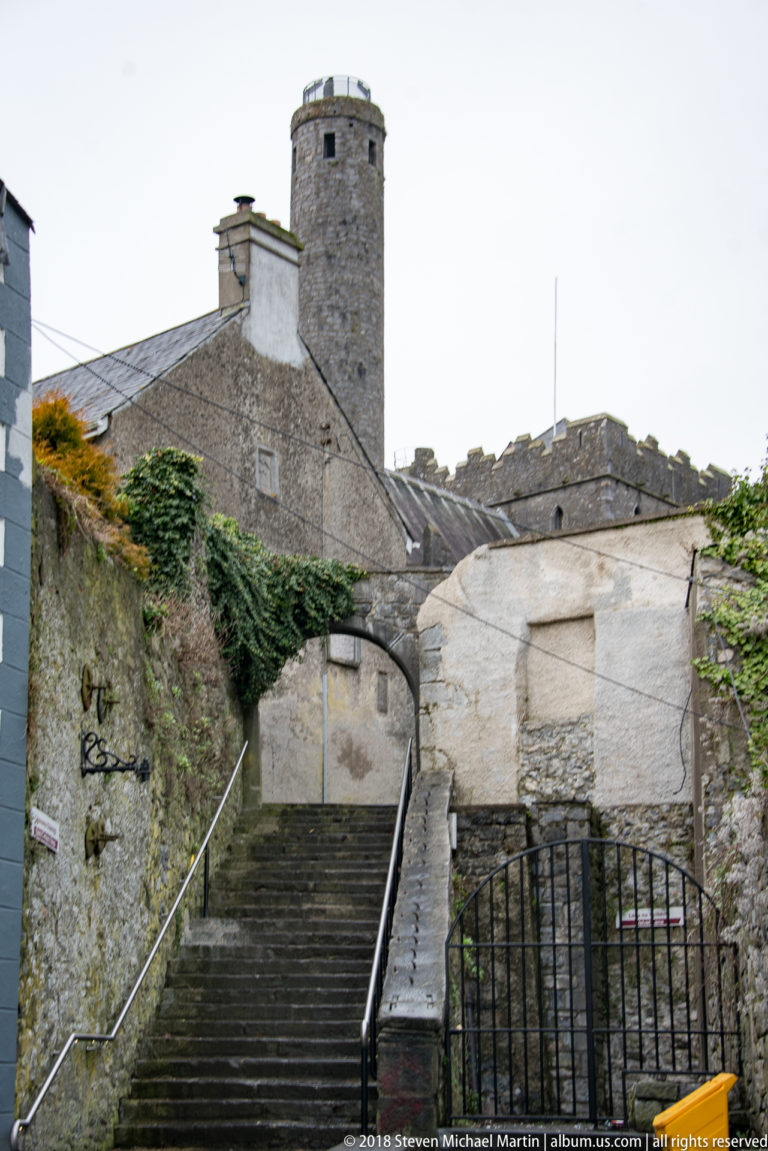 Stone steps and walls lead toward St Canice’s Cathedral, with the tall round tower rising behind the roofline.