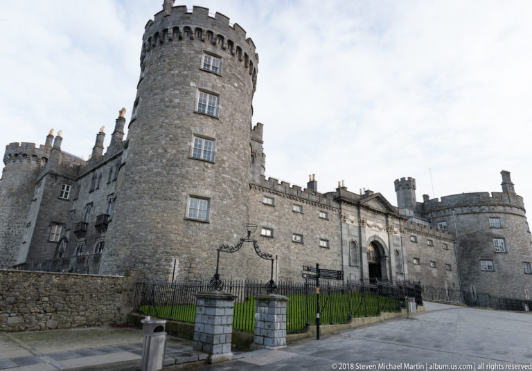 Kilkenny Castle entrance façade with round stone tower, crenellated walls, iron railings, and paved forecourt.