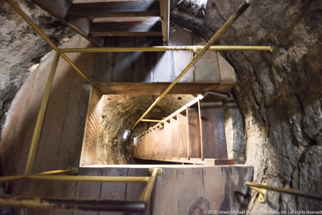 Wooden stair flights turn between platforms inside a narrow, rough-walled stone tower.