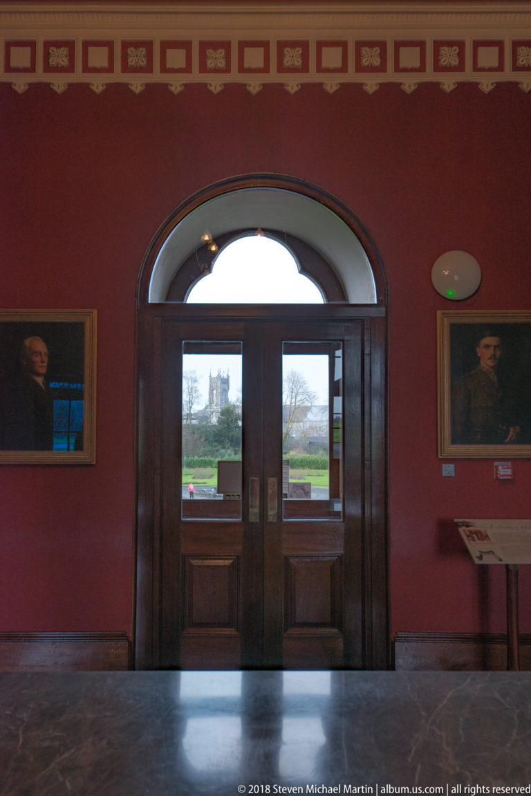Red-walled castle room with portraits flanking a large arched glazed doorway opening toward the grounds.