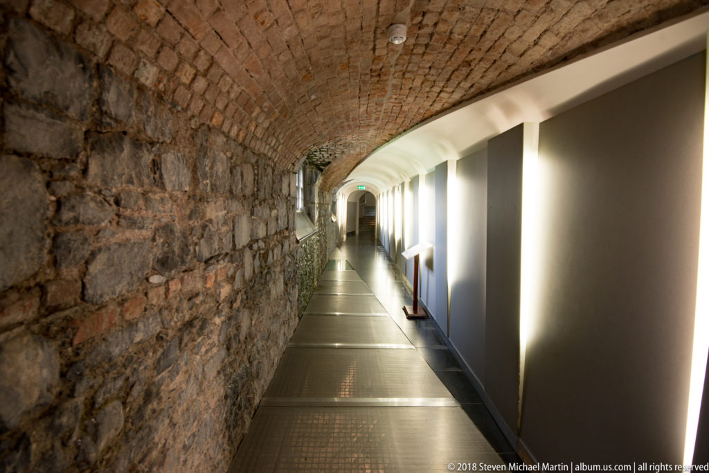 Long castle corridor with brick barrel vault, rough stone wall, modern panels, and floor lighting.