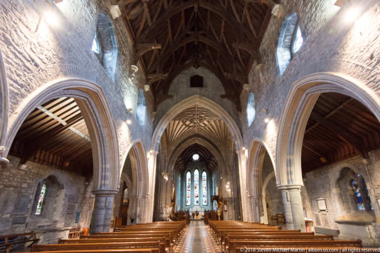 Upward view through St Canice’s Cathedral nave, with pointed stone arches, timber roof and stained-glass windows.
