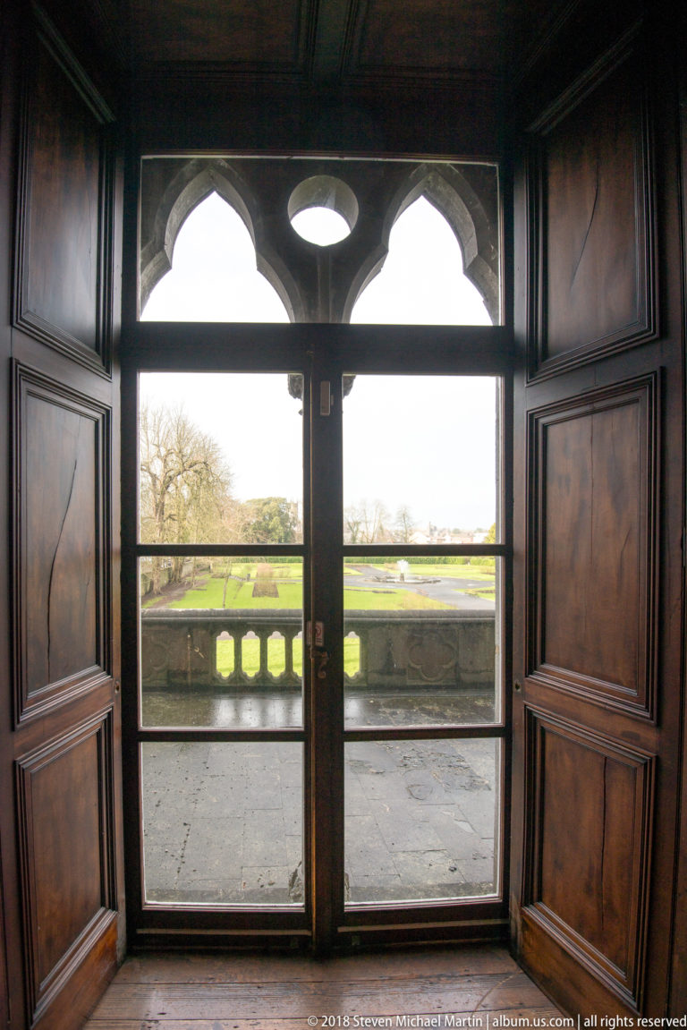 Wood-paneled interior doorway with glass doors opening toward a formal lawn and stone balustrade.
