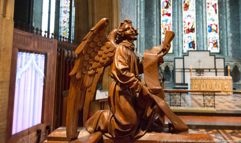 Carved wooden angel with raised hand and feathered wings, with cathedral stained glass and furnishings behind.
