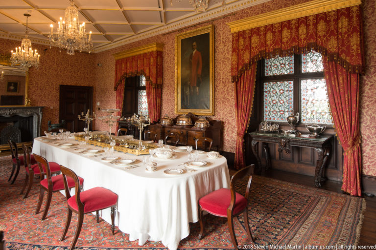 Formal dining room with a long set table, red curtains, patterned walls, portraits, and antique furniture.