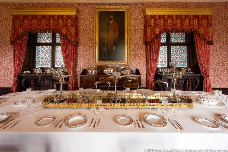 Formal dining table with symmetrical place settings, glassware, silverware, and a long gilt centerpiece beneath a portrait.