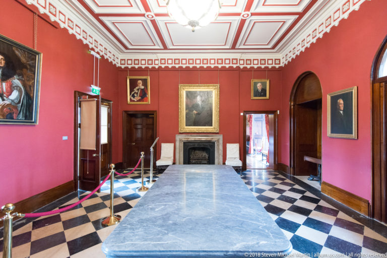 Red castle room with a long central table, black fireplace, portraits, and black-and-white tiled floor.