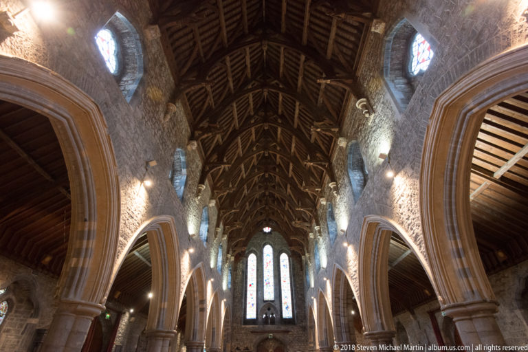 Upward view of dark timber roof trusses above pointed stone arches and small clerestory windows.