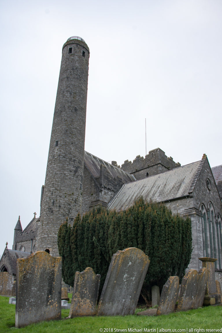 St Canice’s Cathedral round tower rises beside the church above leaning gravestones and clipped shrubs.