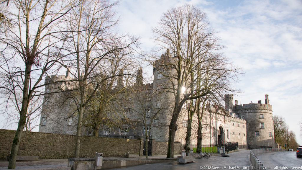 Kilkenny Castle seen behind leafless trees and a stone wall, with multiple towers and rooflines visible.