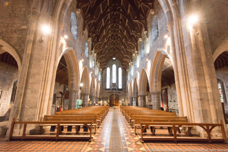 Symmetrical cathedral nave with timber pews, pointed arches, stone piers and patterned central aisle.