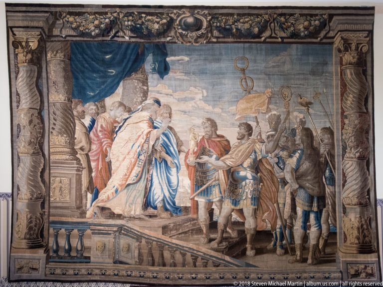 Large tapestry depicting formally dressed figures, armor, drapery, and architectural columns within an ornate border.