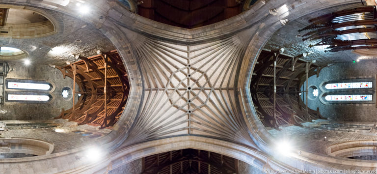Straight-up view of a ribbed stone vault surrounded by arches and dark timber roof sections.