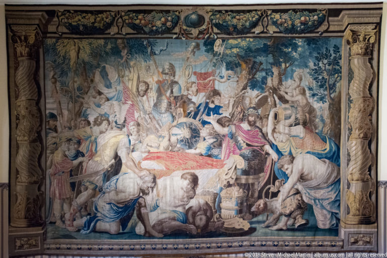 Large tapestry filled with mounted, standing, and kneeling figures, banners, vessels, and ornate border motifs.