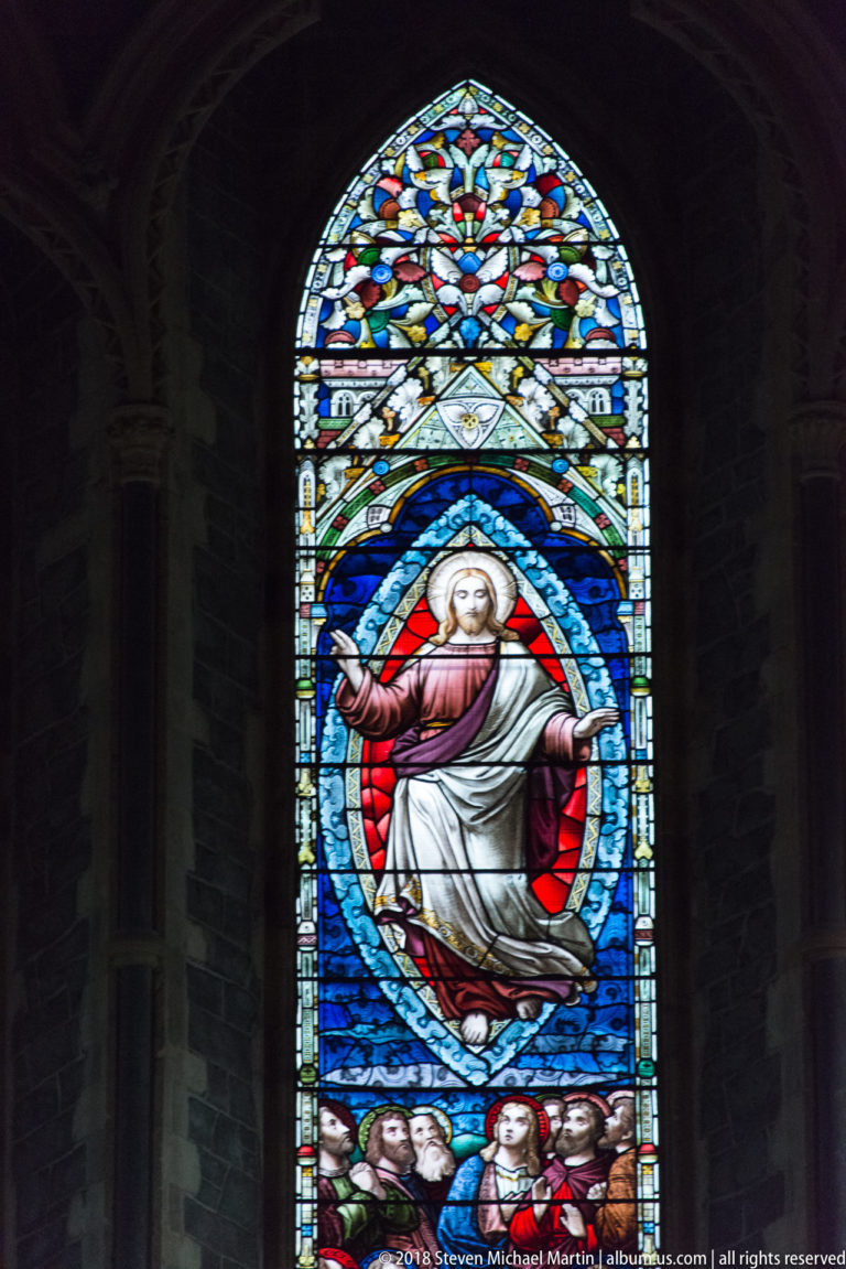 Stained-glass window with a large haloed bearded figure in red and white robes above smaller figures.