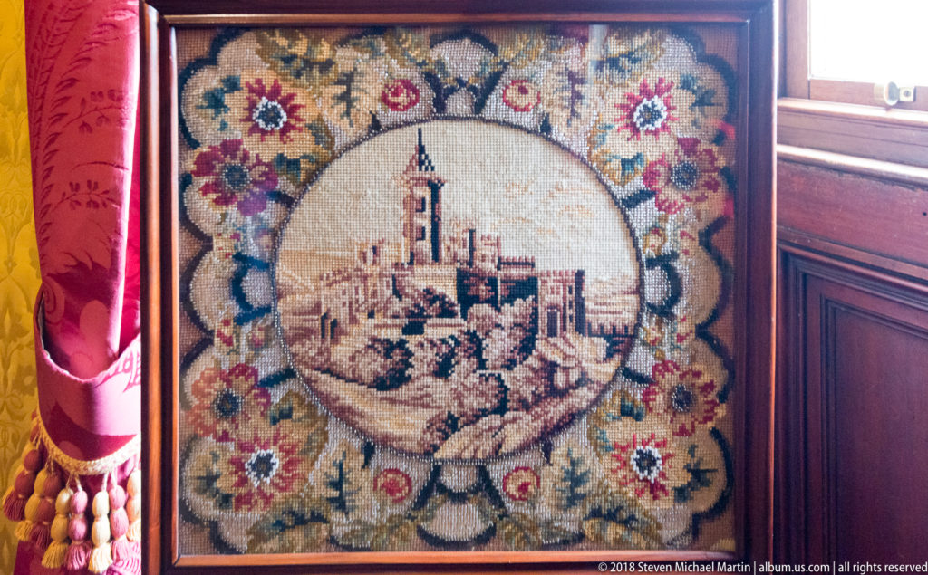 Framed needlework showing a turreted castle inside a circular medallion surrounded by colorful flowers.