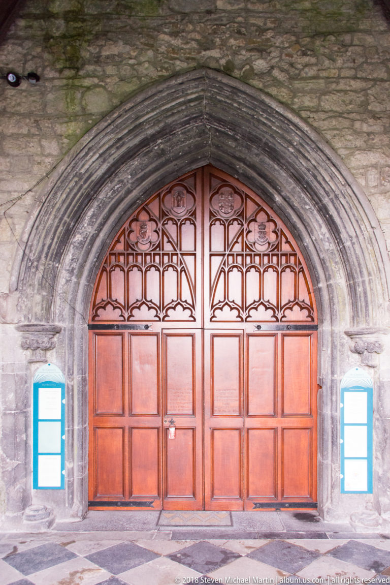 Paired reddish wooden doors beneath a pointed stone arch and glazed tracery inside the cathedral.