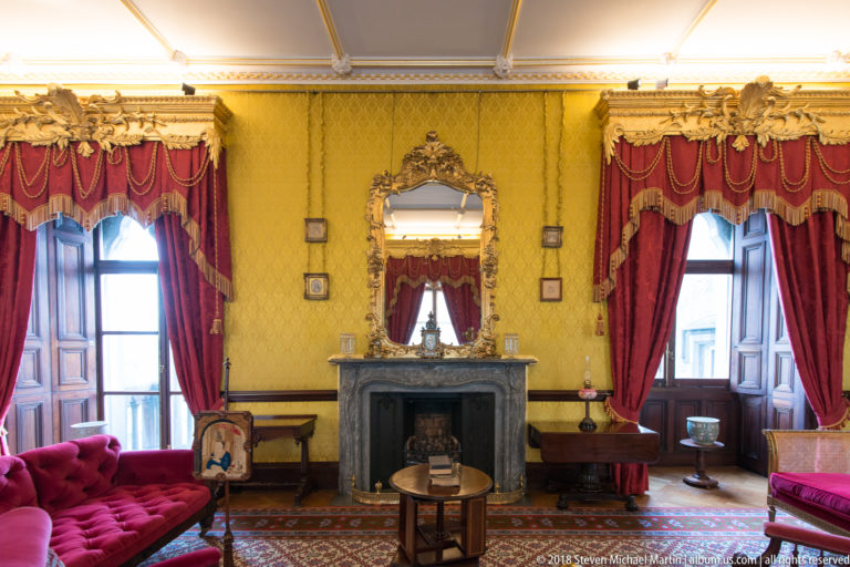 Yellow drawing room with red curtains, gilded mirror, black fireplace, upholstered sofas, and small tables.