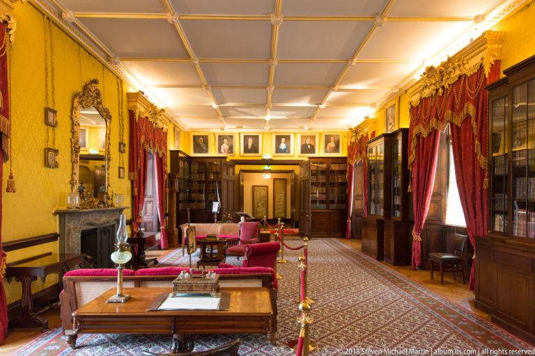 Long historic library with bookcases, tables, red curtains, patterned carpet, and portraits at the far end.