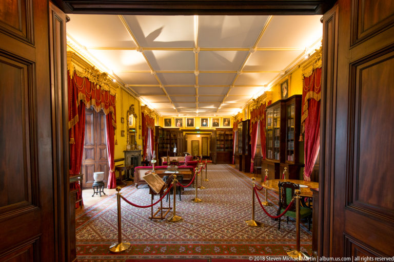 Kilkenny Castle library seen through a dark wooden doorway, with bookcases, red curtains, carpet, and furniture beyond.
