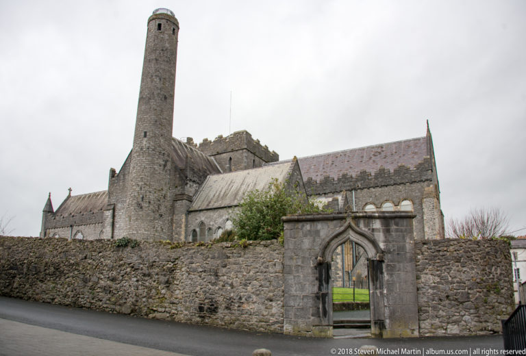 St Canice’s Cathedral and round tower seen beyond a stone wall and pointed gateway from the street.