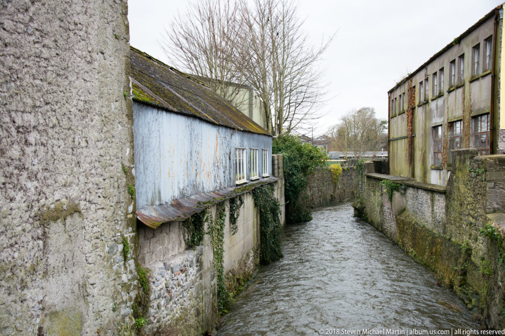 Narrow watercourse flowing between weathered stone walls and older riverside buildings in Kilkenny.