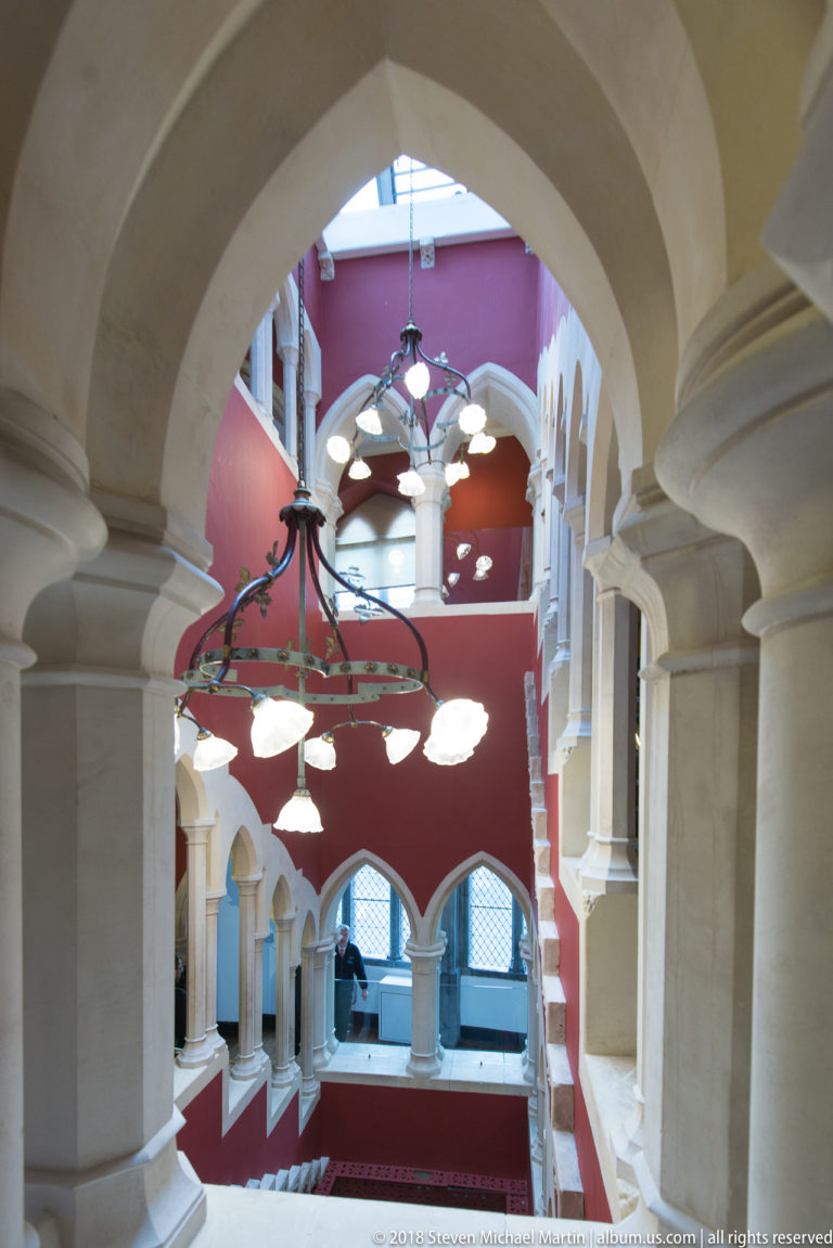Multi-level castle stair hall with pale stone arches, red walls, windows, and hanging globe light fixtures.