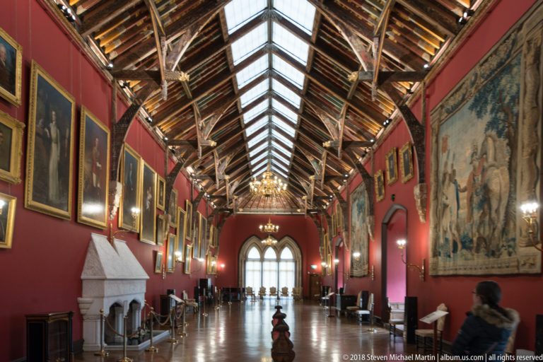 Long red-walled picture gallery with portraits, tapestries, timber roof, skylight, fireplace, and polished floor.