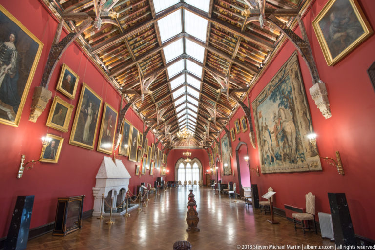 Wide-angle interior of Kilkenny Castle’s Picture Gallery with red walls, paintings, tapestries, skylit timber roof, and fireplace.