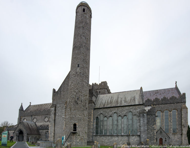 Full-height stone round tower beside the gabled and buttressed exterior of St Canice’s Cathedral.