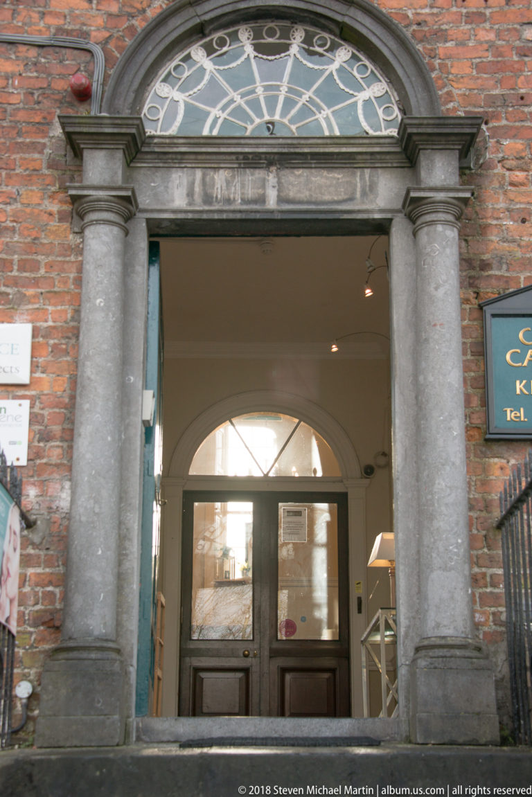Red-brick wall with a classical stone doorway, paired columns, and a semicircular fanlight.