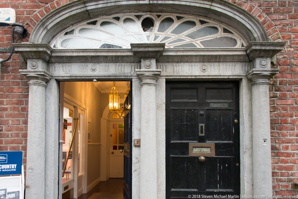 Red-brick façade with a classical stone doorway, semicircular fanlight, paired black doors and lit hall beyond.