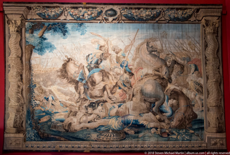 Large tapestry showing mounted combat with horses, armed figures, and fallen bodies within an ornate border.