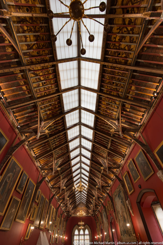 Upward view along Kilkenny Castle’s Picture Gallery, with painted timber roof, central skylight, pendant lights, red walls, and framed portraits.