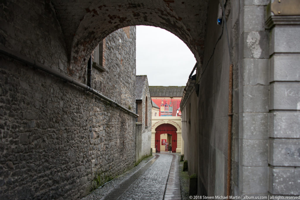 Cobblestone lane running beneath a dark stone arch between high masonry walls toward a red doorway.