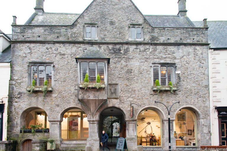 Rough stone building with ground-floor arches, flower-filled windows, and a small projecting oriel window above the arcade.