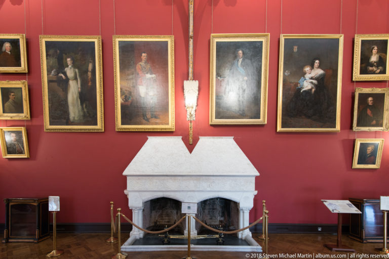 Red gallery wall filled with gilt-framed portraits above a pale stone fireplace, with ropes and small interpretive stands in front.
