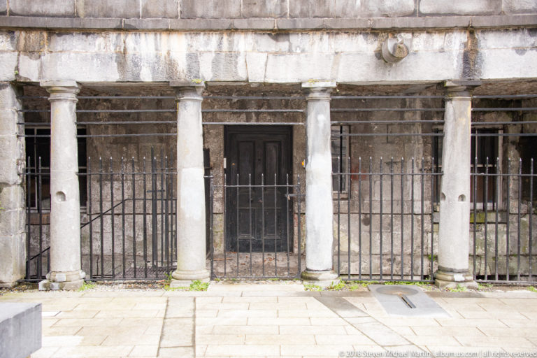 Row of pale stone columns and shadowed openings behind tall black iron railings on a paved street.