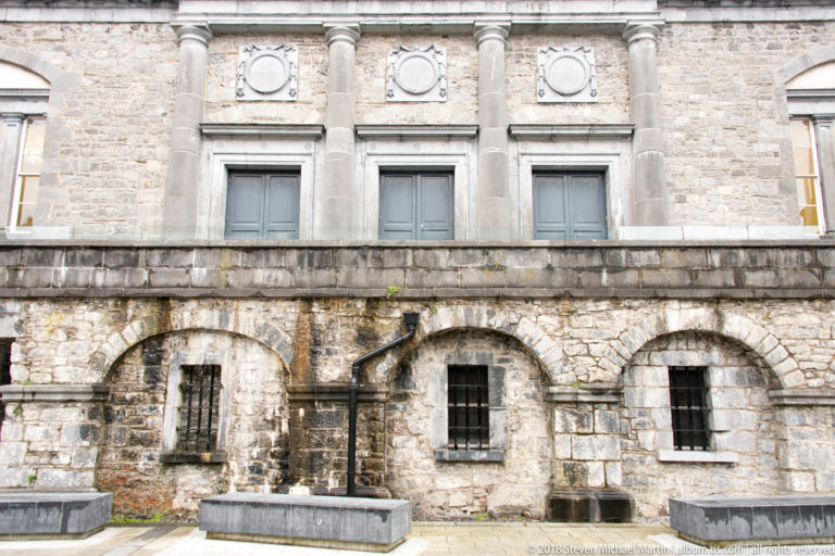 Historic stone façade with three barred arched windows below rectangular upper windows separated by classical pilasters.