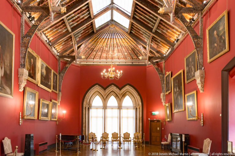 End of the Picture Gallery with a three-part pointed window, chandelier, chairs, red walls, portraits, and faceted painted timber roof.