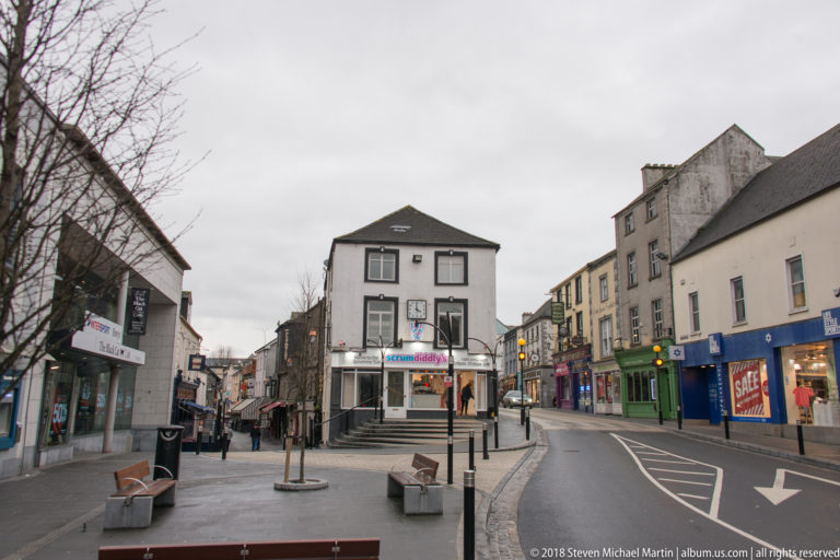 Curving town street with shops, benches, traffic lights, road markings, and mixed historic buildings under an overcast sky.