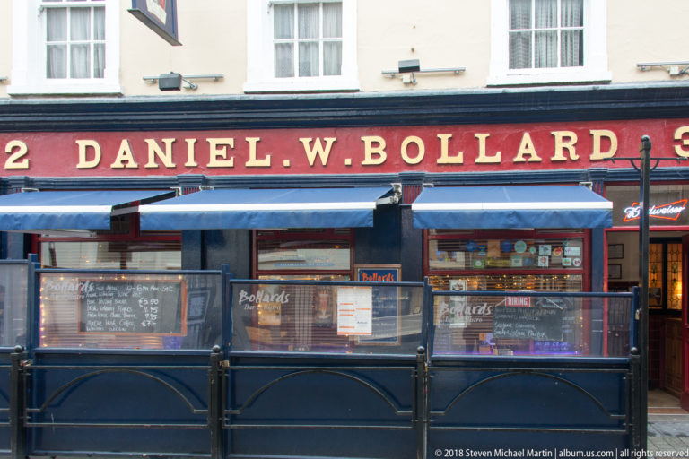 Daniel W. Bollard pub frontage with red signboard, gold lettering, blue awnings, and enclosed outdoor seating.