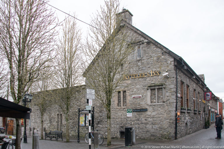 Gabled stone exterior of Kyteler’s Inn with small windows, wall lettering, outdoor tables, bicycles, and street signs.