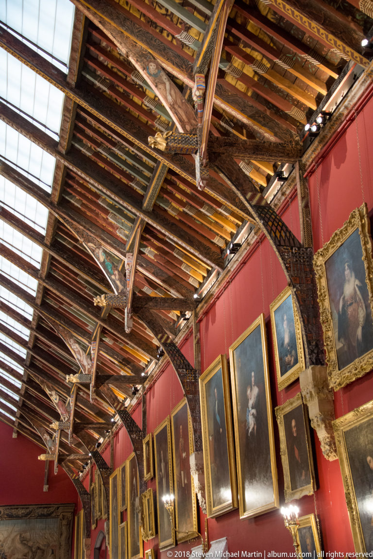 Diagonal view of painted timber roof beams above red walls hung with large gilt-framed portraits in the Picture Gallery.